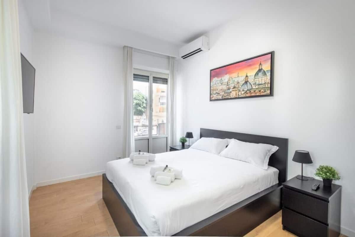 Una habitación de hotel con cama, mesita de noche, un cuadro y ventana.
