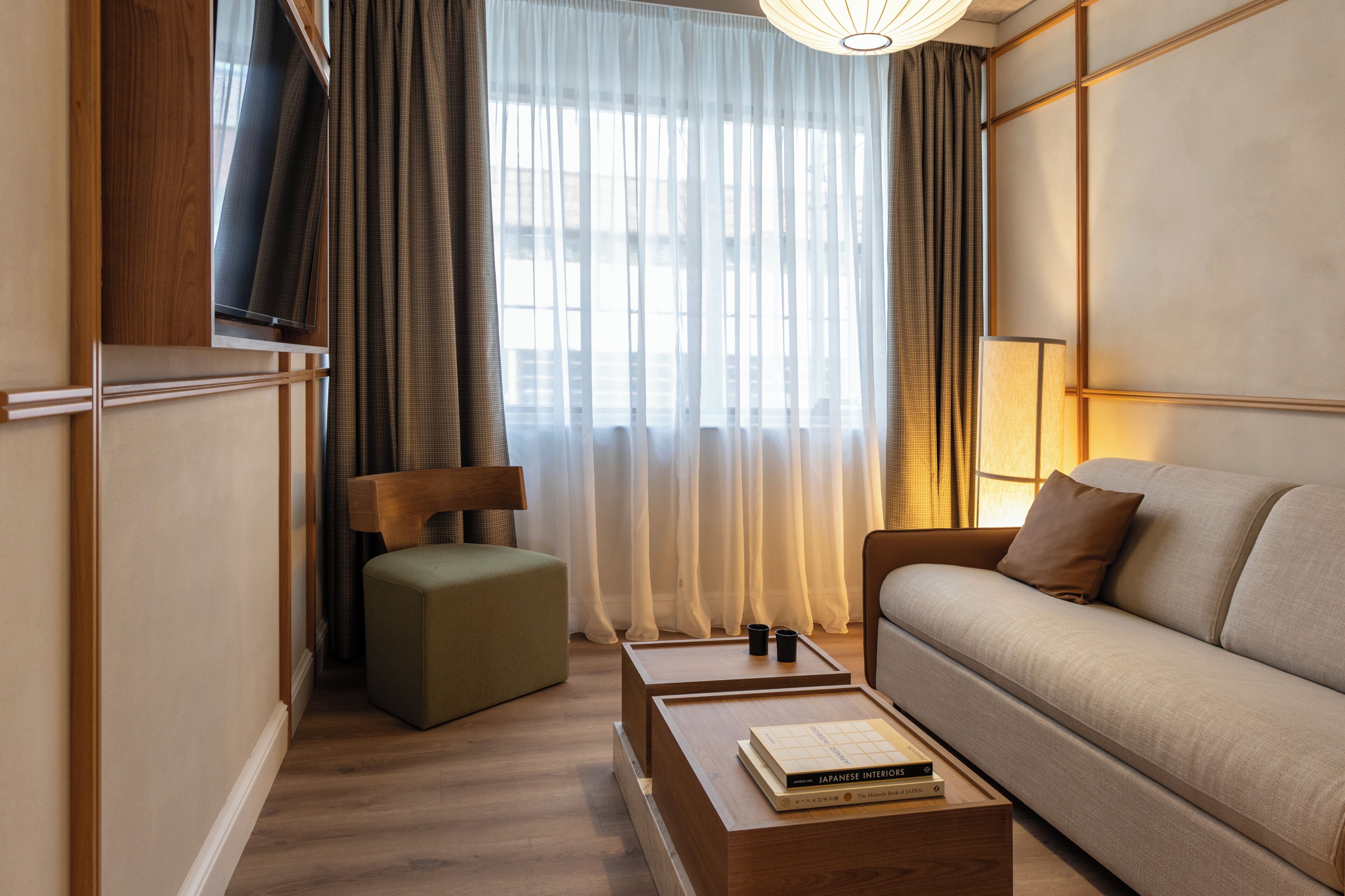 A modern hotel room with a sofa, a small table with books, a wooden bench, and a large window with curtains.
