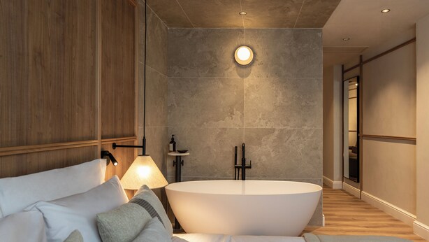 Moderni hotellihuone, jossa on suuri kylpyamme, puinen sängynpääty ja yöpöytä, jossa on lamppu.