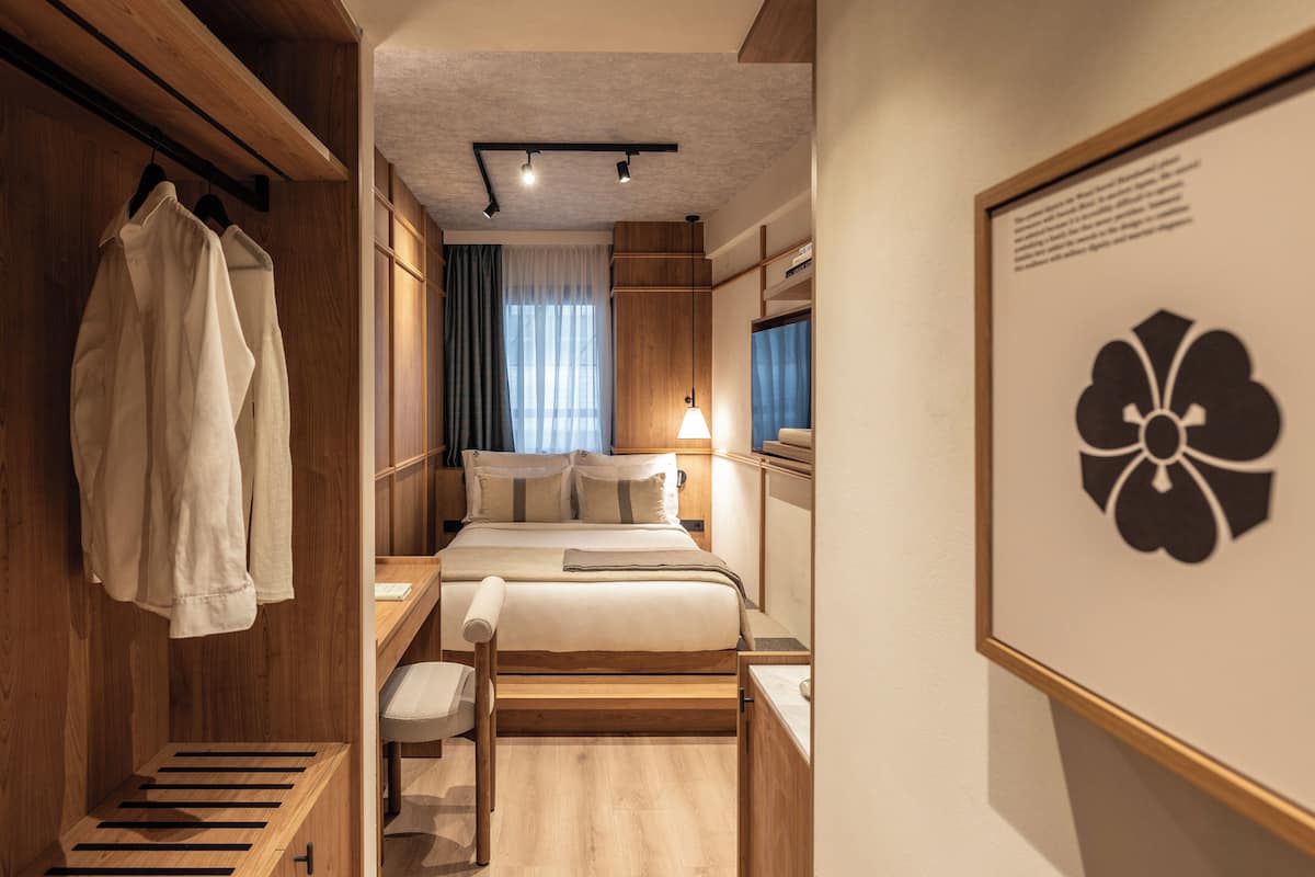 A hotel room with a wooden wardrobe, a bed with a headboard, a bedside table, a chair, and a framed picture on the wall.
