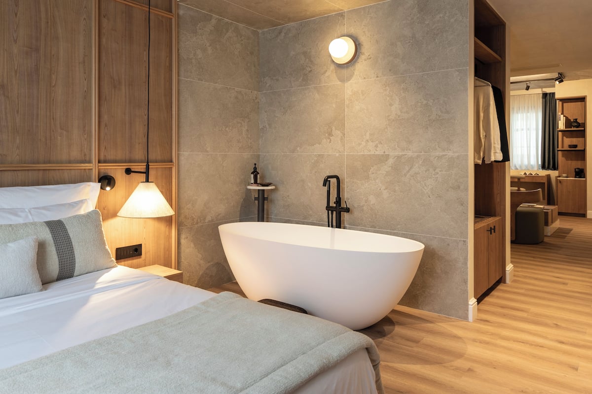 A modern hotel room with a large bathtub, a wooden headboard, a bedside table with a lamp, and a view into the bathroom.