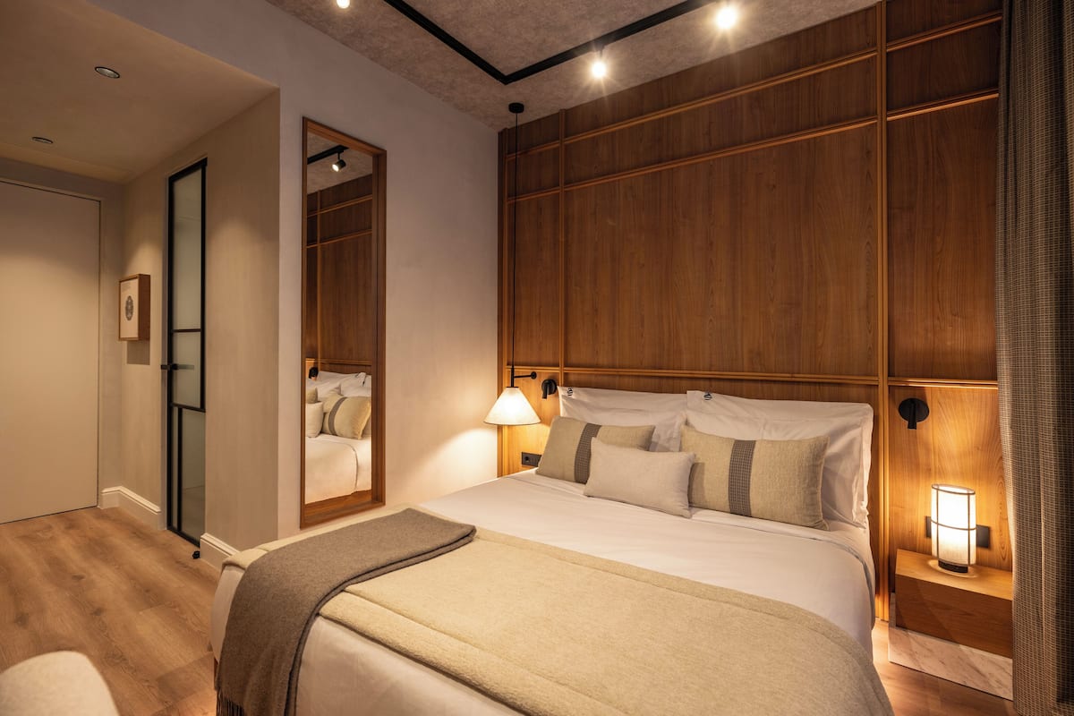 A hotel room with a large bed, wooden paneling, a nightstand with a lamp, a mirror, and a door leading to another room.