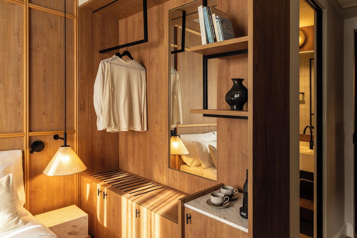 A hotel room with wooden paneling, a bed, a bedside table with a lamp, a mirror, a shelf with books, and a small table with cups and a vase.
