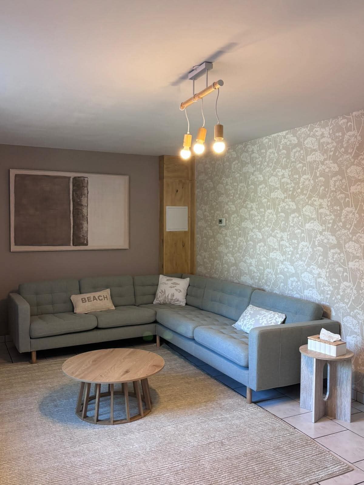 A living room with a sectional sofa, a round wooden coffee table, a small side table, and a patterned wallpaper.