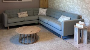 Ein Wohnzimmer mit einem Ecksofa, einem runden Holztisch, einem kleinen Beistelltisch und einer Wand mit Musterung.