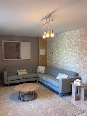 Ein Wohnzimmer mit einem Ecksofa, einem runden Holztisch, einem kleinen Beistelltisch und einer Wand mit Musterung.