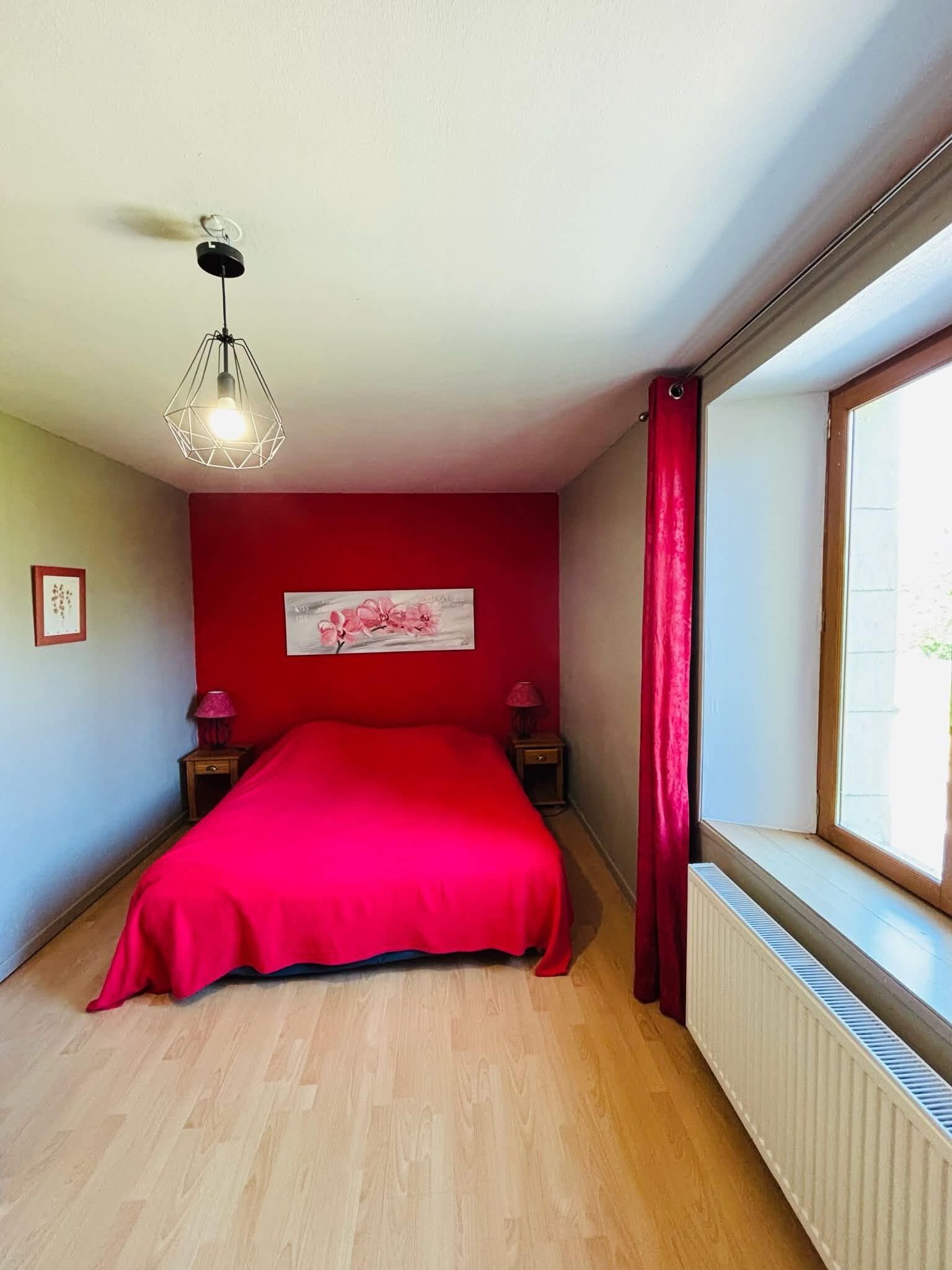 Een slaapkamer met een rode wand, een bed met een rode dekbedovertrek, een raam met uitzicht, een radiator en een hanglamp.