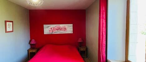 Een slaapkamer met een rode wand, een bed met een rode dekbedovertrek, een raam met uitzicht, een radiator en een hanglamp.