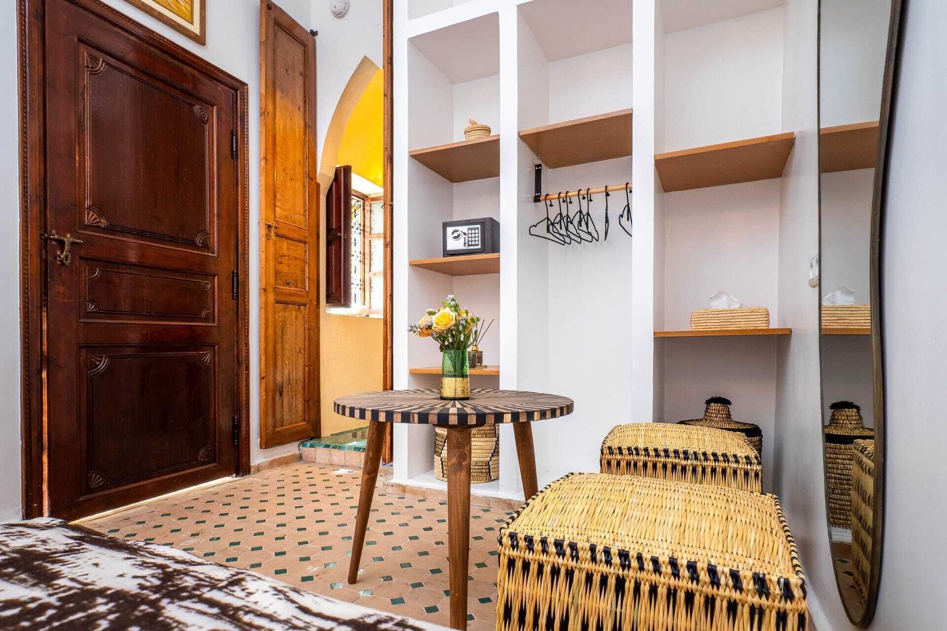 A room with a large wooden door, a round table with a vase of flowers, a woven bench, and white shelving units.