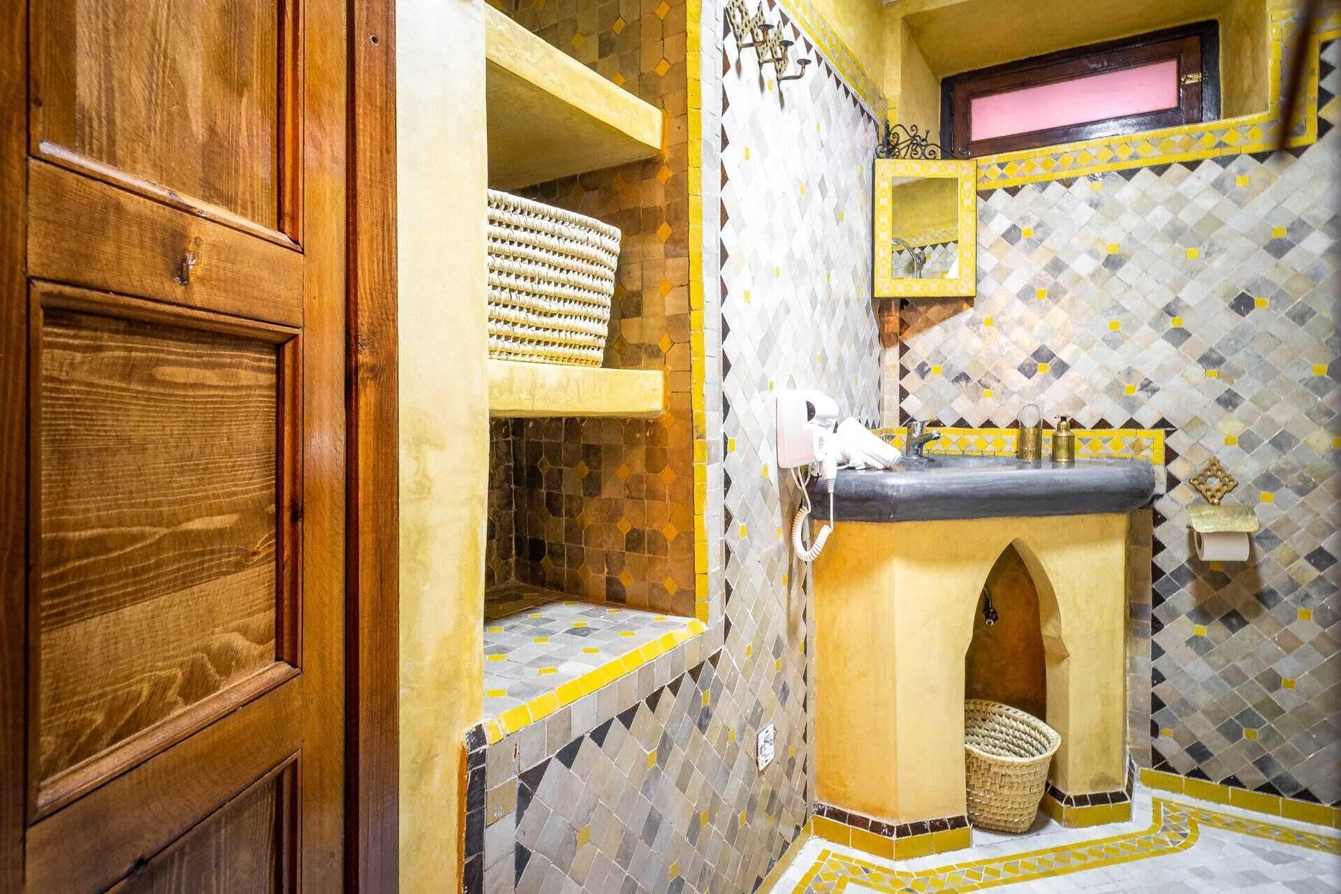 A bathroom with a wooden cabinet, a shelf with folded towels, a sink with a mirror, and a patterned tile wall.