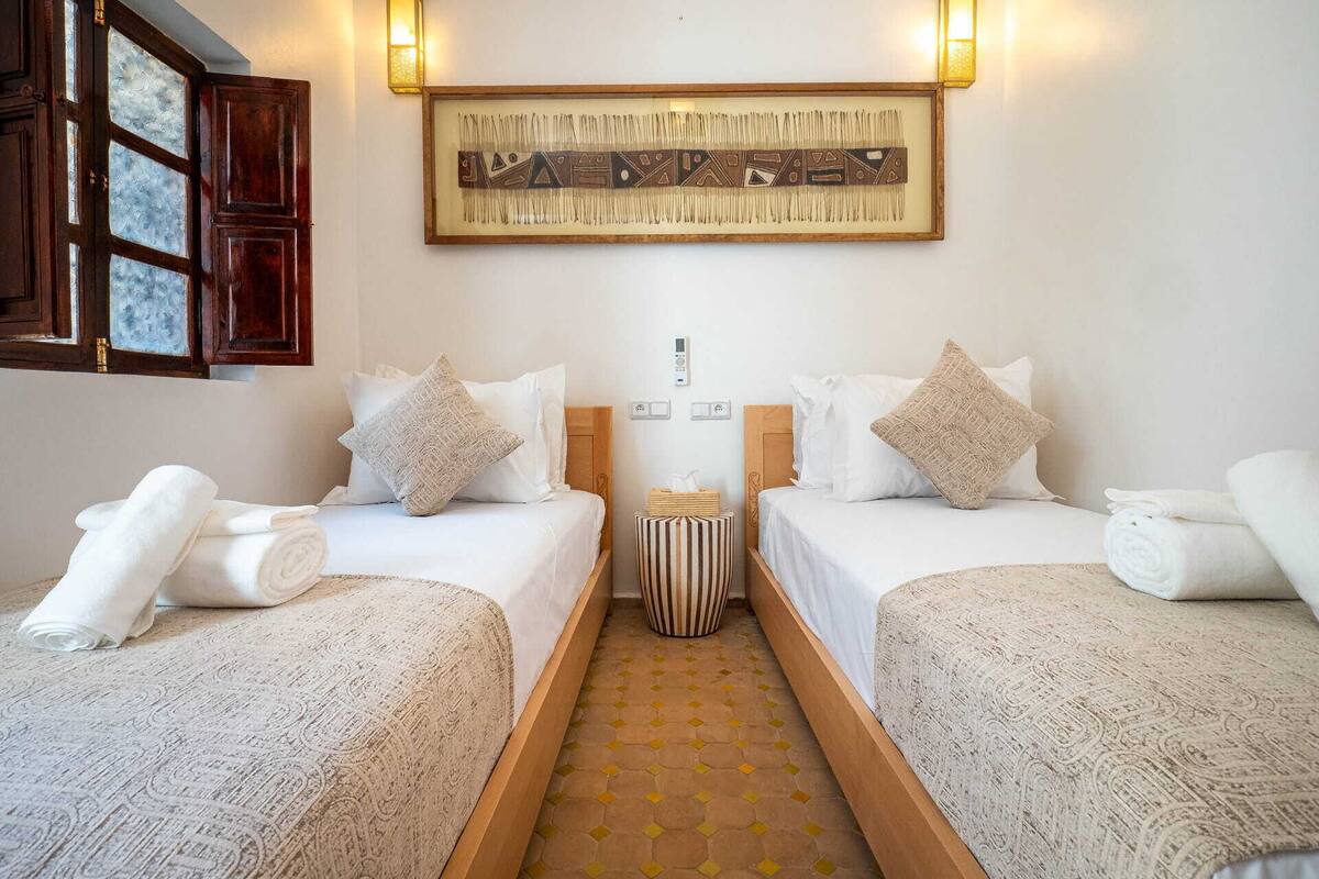 A hotel room with two beds, a small side table, a framed artwork above the beds, and a window with a view.