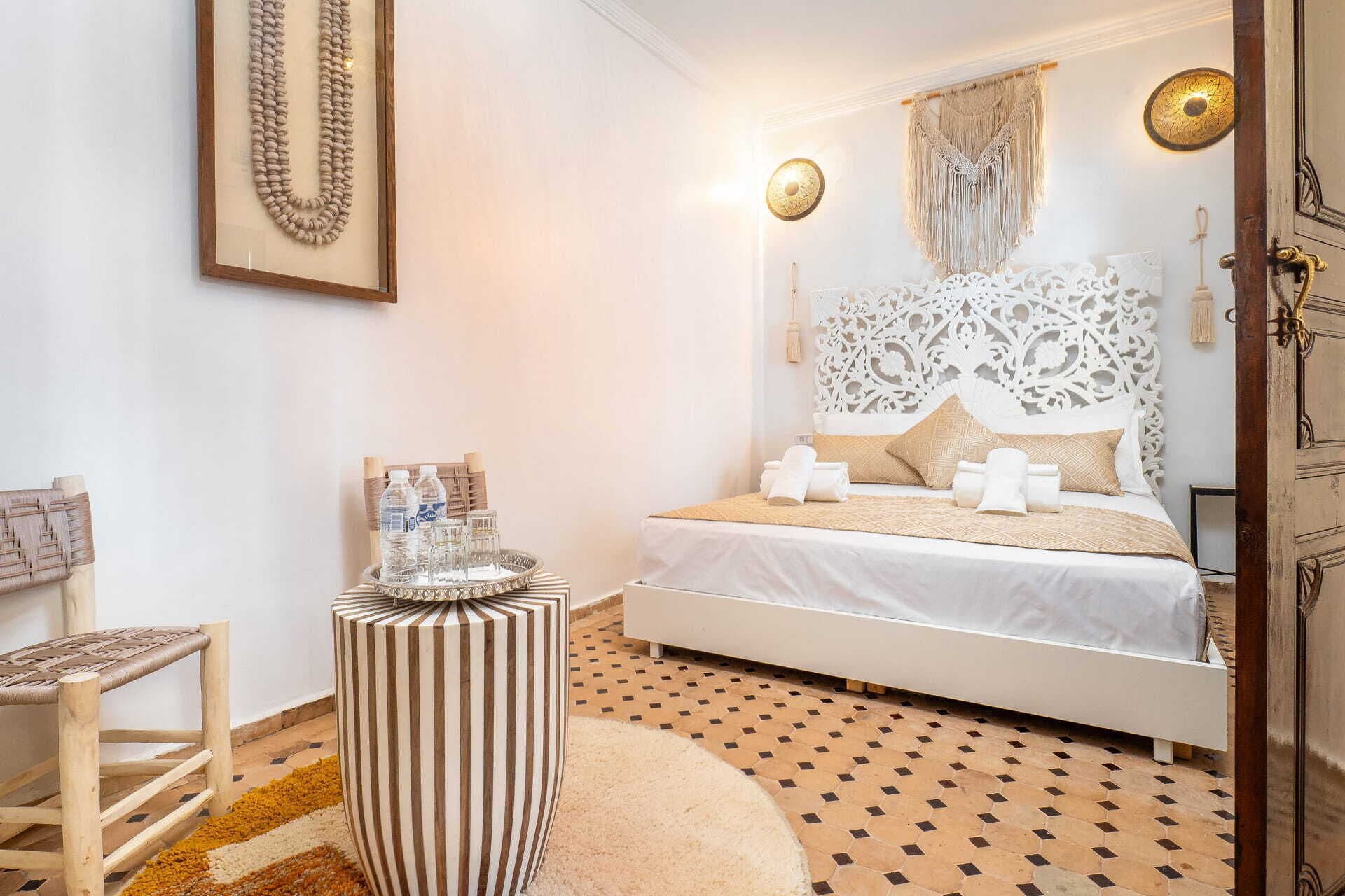 A bedroom with a bed, a side table, a chair, a macrame wall hanging, and a patterned floor.