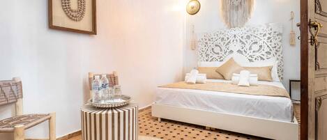 A bedroom with a bed, a side table, a chair, a macrame wall hanging, and a patterned floor.