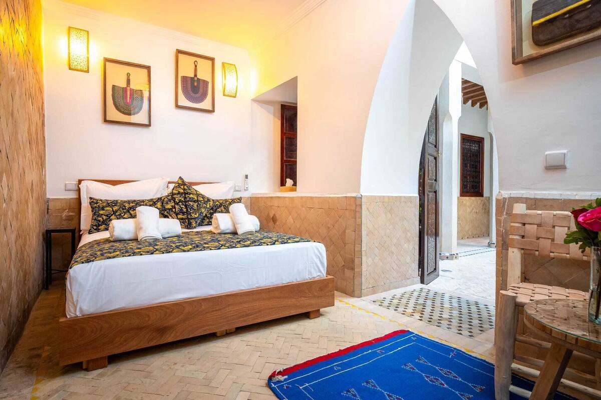 A bedroom with a wooden bed, white linens, patterned pillows, a blue and red rug, and wall-mounted lights.