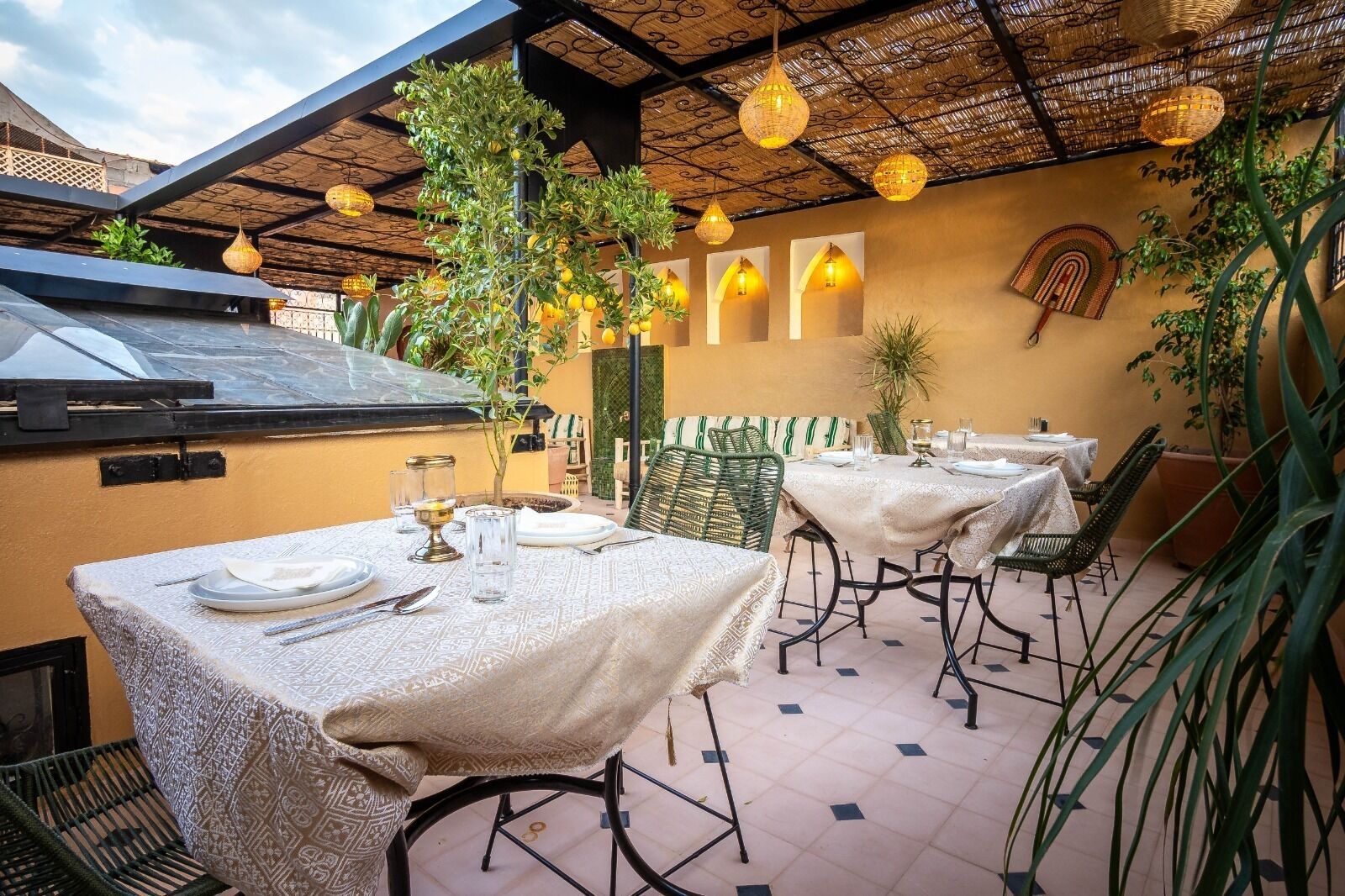 An outdoor dining area with tables set for two, covered with patterned tablecloths, surrounded by chairs, and decorated with hanging lamps.
