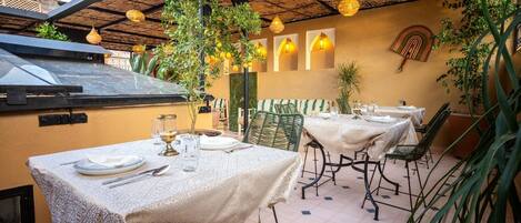 Un espace de restauration en plein air avec des tables pour deux, recouvertes de nappes à motifs, entourées de chaises et décorées de lampes suspendues.