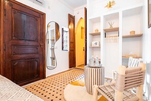 A hotel room with a large wooden door, a mirror, a small round table with a striped chair, and shelves with various items. - Riad Koutchi (Marrakech)