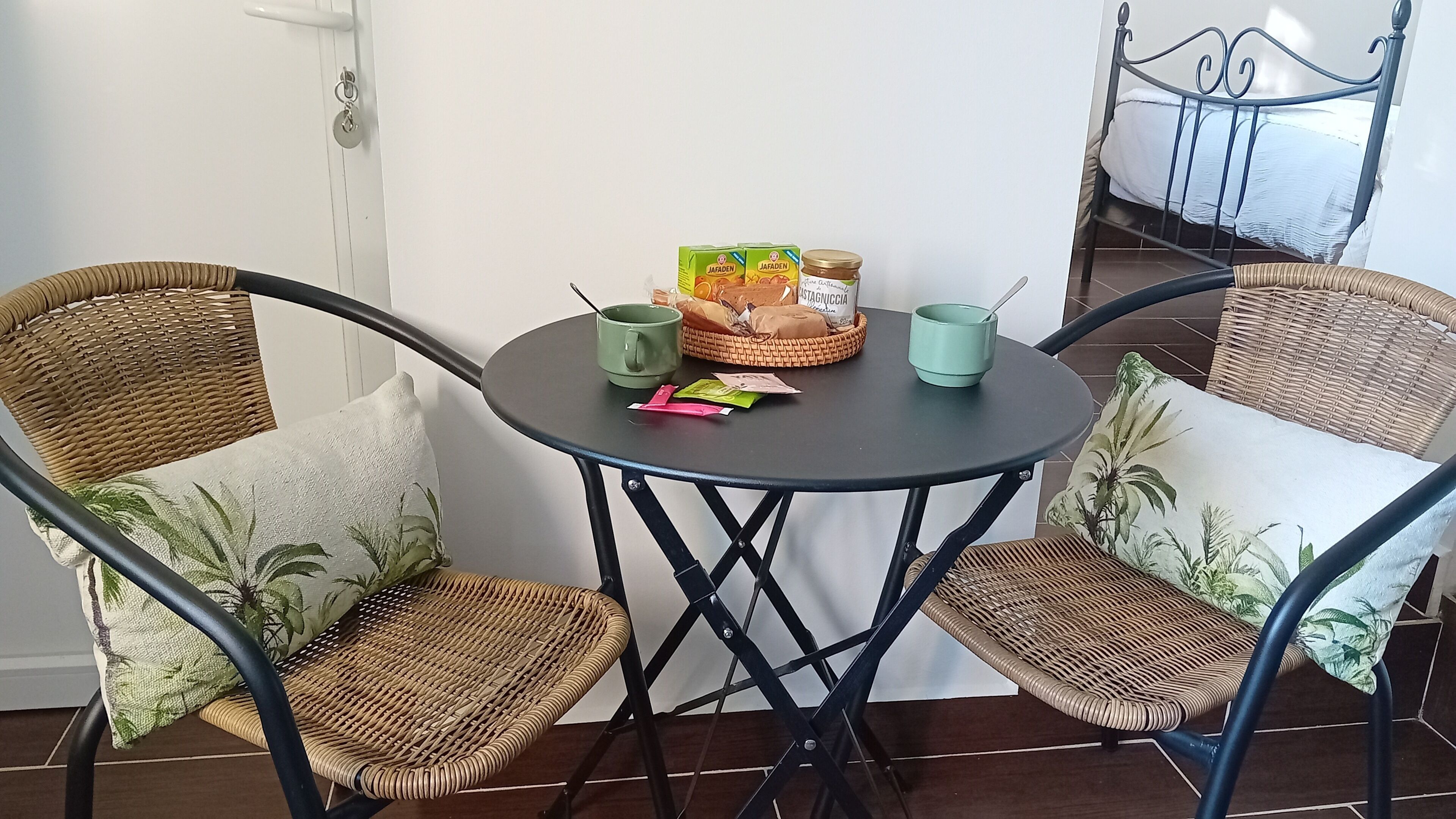 Et lille rundt bord med fletstole, en kurv med brød, et syltetøjsglas og en grøn kop.