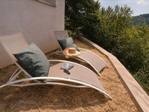 Eine Liege mit Kissen, Hut und Buch auf einem Balkon mit Blick auf Bäume und Hügel.
