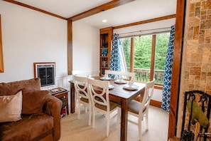 Uma sala de estar acolhedora com um sofá marrom, uma mesa de jantar de madeira com cadeiras brancas, uma estante de livros e uma janela com cortinas.