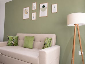 En beige sofa med grønne blomstermønstrede puder, en gulvlampe og fire indrammede kunstværker på en grøn væg.