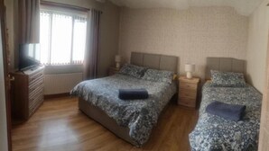 A bedroom with two beds, a dresser, a radiator, and a window with curtains. - Church View  (Ballycastle)