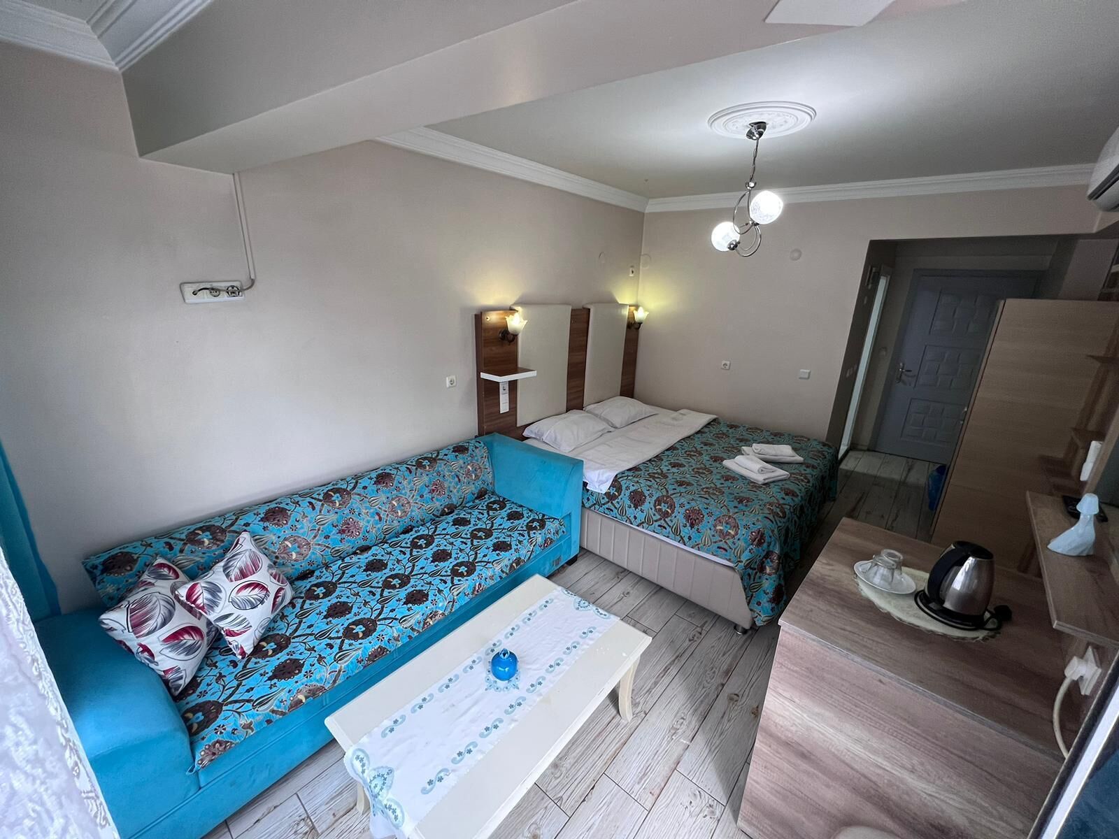 A small, well-lit room with a sofa, a bed, a bedside table, and a mirror.