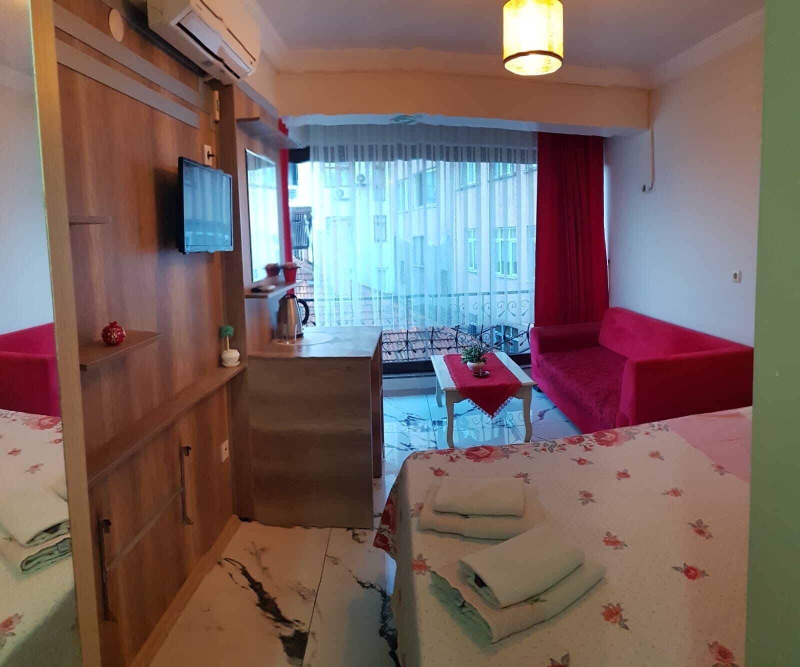 A hotel room with a balcony, a sofa, a small table with a red tablecloth, and a TV mounted on the wall.