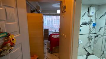 A hallway with a door leading to a room with a bed and a red blanket, a shower area with a glass door, and a ceiling light.