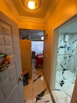 A hallway with a door leading to a room with a bed and a red blanket, a shower area with a glass door, and a ceiling light.