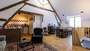 A loft-style room with exposed wooden beams, a kitchen area, a dining table with chairs, and a staircase.
