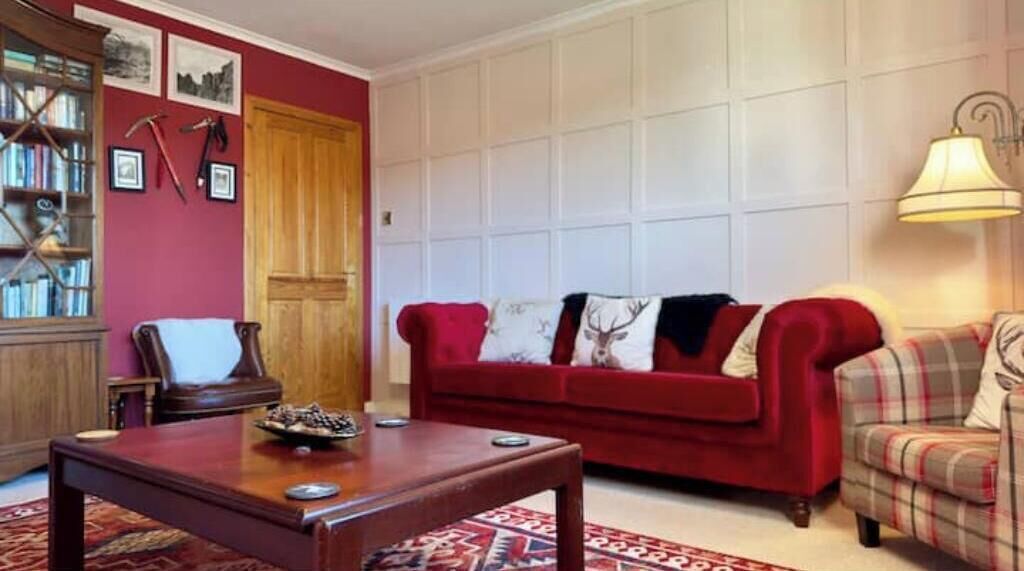 A red sofa, a plaid armchair, a wooden table, and a bookshelf with books and pictures.