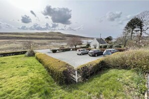 Ein Parkplatz mit Autos, ein Gebäude mit Satteldach und eine hügelige Grünlandschaft im Hintergrund.