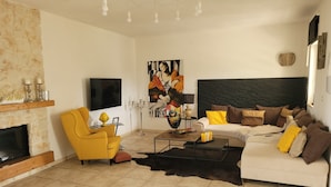 A living room with a fireplace, a yellow armchair, a white sofa with brown cushions, a large abstract painting, and a flat-screen TV mounted on the wall.