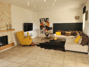 Una sala de estar con chimenea, un sillón amarillo, un sofá blanco con cojines marrones, un cuadro abstracto grande y un televisor de pantalla plana montado en la pared.