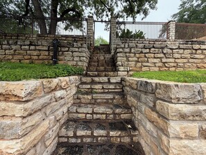 Eine Steintreppe führt zu einem von einem Metallzaun umgebenen Bereich, im Hintergrund sind Bäume zu sehen.