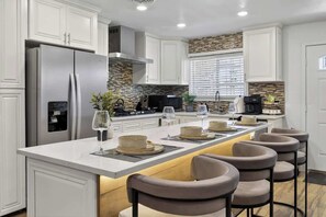 A modern kitchen with white cabinetry, a central island with bar stools, a stainless steel refrigerator, and a built-in oven. - 7BR Newly Remodeled Home w/ Arcades, Sleeps 20 (North Las Vegas)