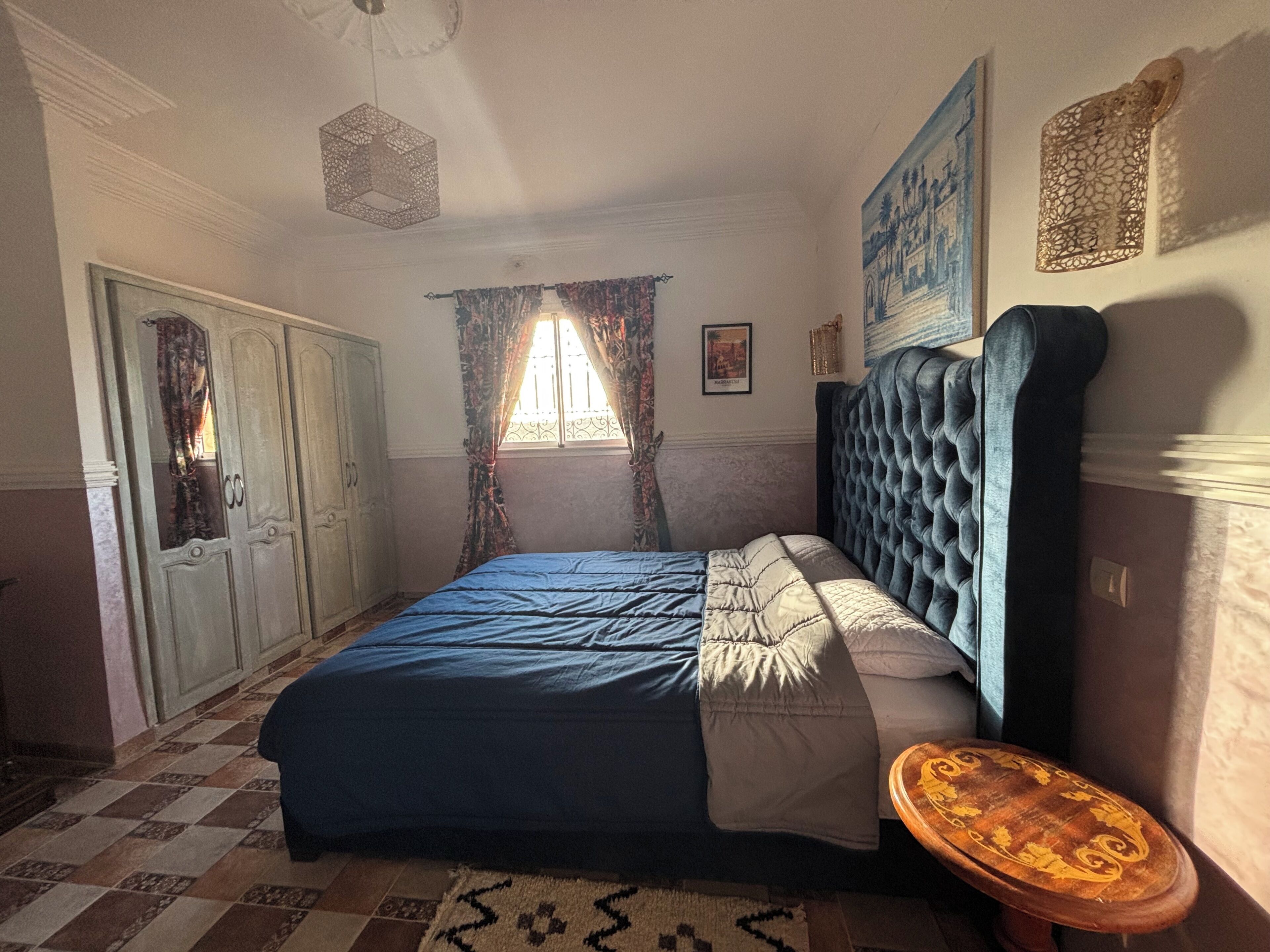 Un dormitorio con cama, armario, ventana con cortinas, una obra de arte en la pared y una lámpara en el techo.