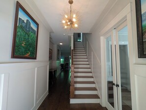 A hallway with a staircase, a chandelier, and framed artwork on the walls.