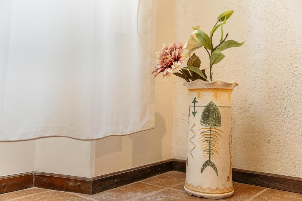 Un angolo con una tenda bianca, un pavimento piastrellato e un vaso decorato con un motivo a pesce e una pianta.