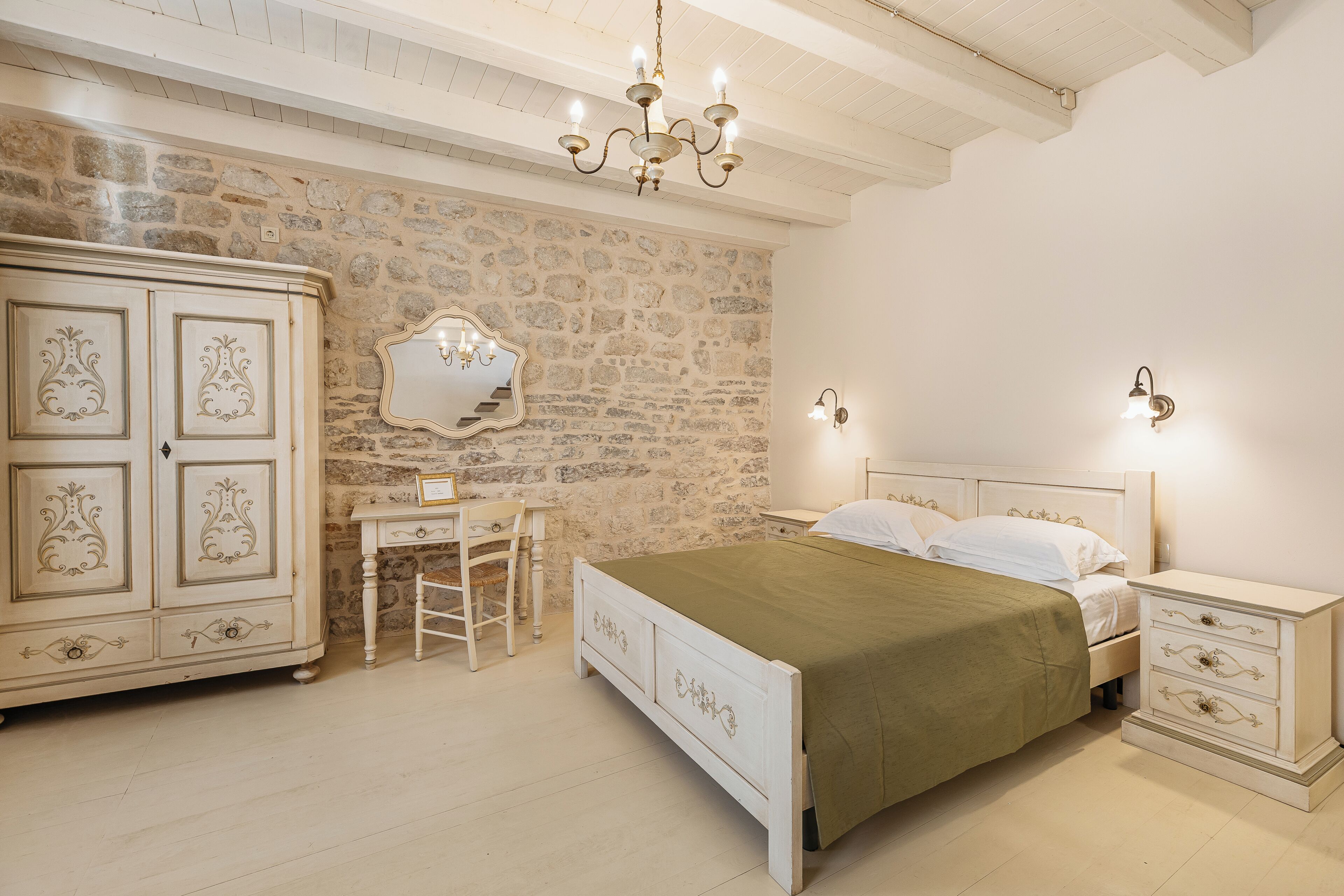 A bedroom with a stone wall, a bed with a green bedspread, a desk with a chair, and a wardrobe.