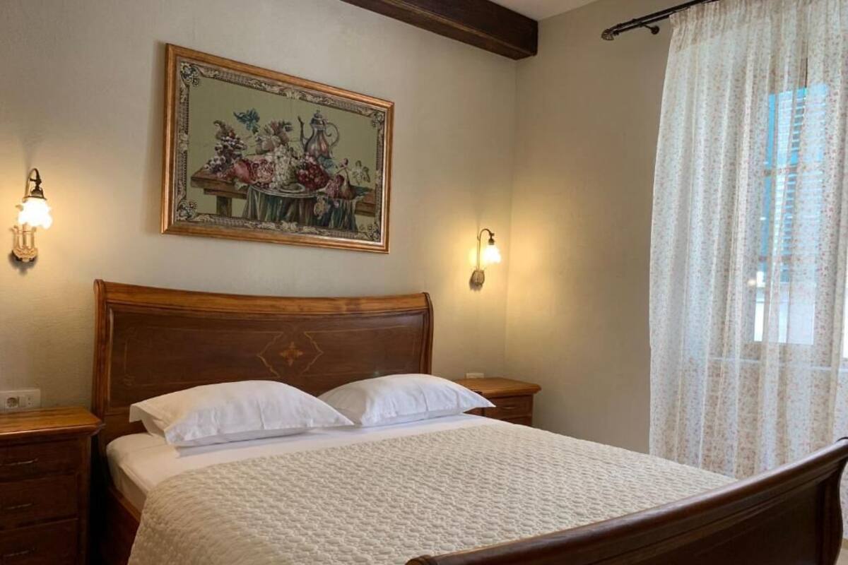 Una cama bien hecha con cabecera de madera, dos mesitas de noche, un cuadro colgado en la pared y una ventana con cortinas traslúcidas.