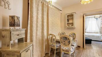 Um quarto com piso de madeira, uma mesa de jantar com cadeiras, uma cama com dossel, um espelho e um lustre.