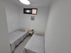 Ein kleines, sauberes Zimmer mit zwei Einzelbetten, einem Nachttisch mit Lampe, einem kleinen Fenster und einem gerahmten Bild an der Wand.
