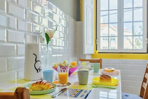 Una mesa de desayuno con diversos platos, un florero con una flor y una ventana con vistas a edificios.
