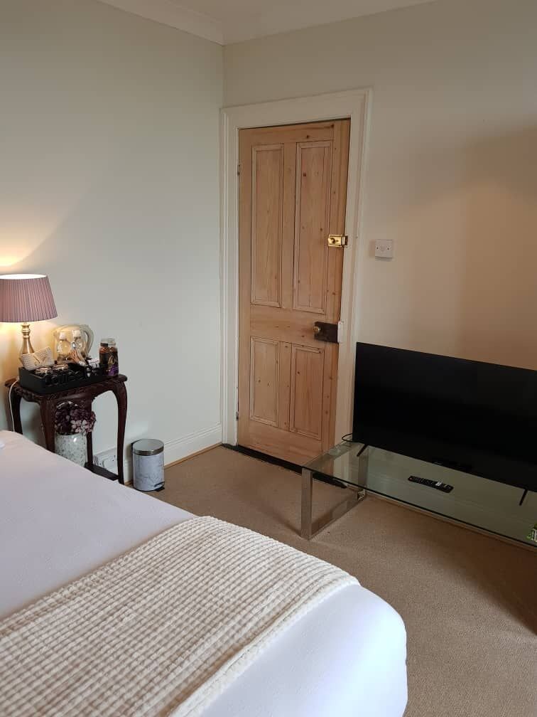 Una camera d'albergo con un letto, una televisione su un tavolino in vetro, una porta in legno e un piccolo tavolo con una lampada e oggetti decorativi.