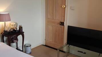 Una camera d'albergo con un letto, una televisione su un tavolino in vetro, una porta in legno e un piccolo tavolo con una lampada e oggetti decorativi.