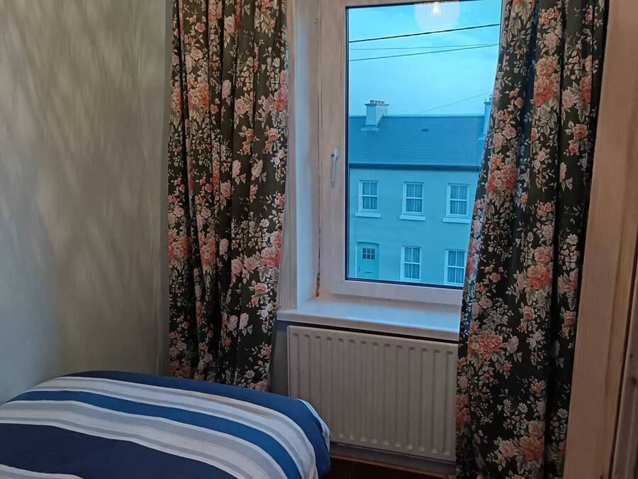 Quarto com cama, janela com cortinas florais, radiador e vista para um edifício.
