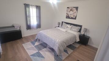 A bedroom with a bed, bedside tables, a nightstand, a window with curtains, and a wall-mounted flower painting.