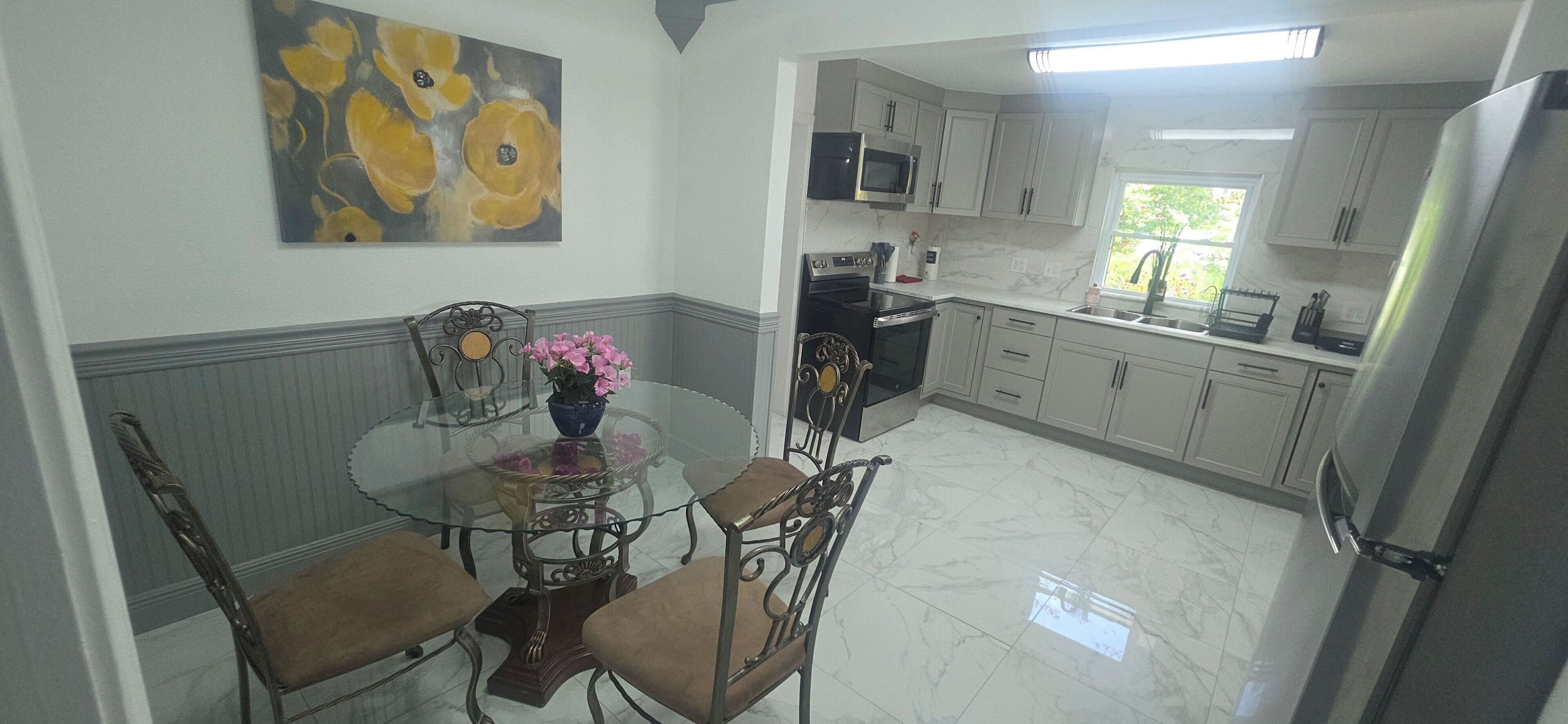 A kitchen with white cabinets, a glass dining table with chairs, a painting on the wall, and a window with a view.